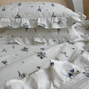 May include: White bedding set with a floral pattern in blue and beige. The set includes two ruffled pillowcases and a sheet, all with a textured, waffle-like weave. The floral design adds a touch of elegance to the bedroom decor.