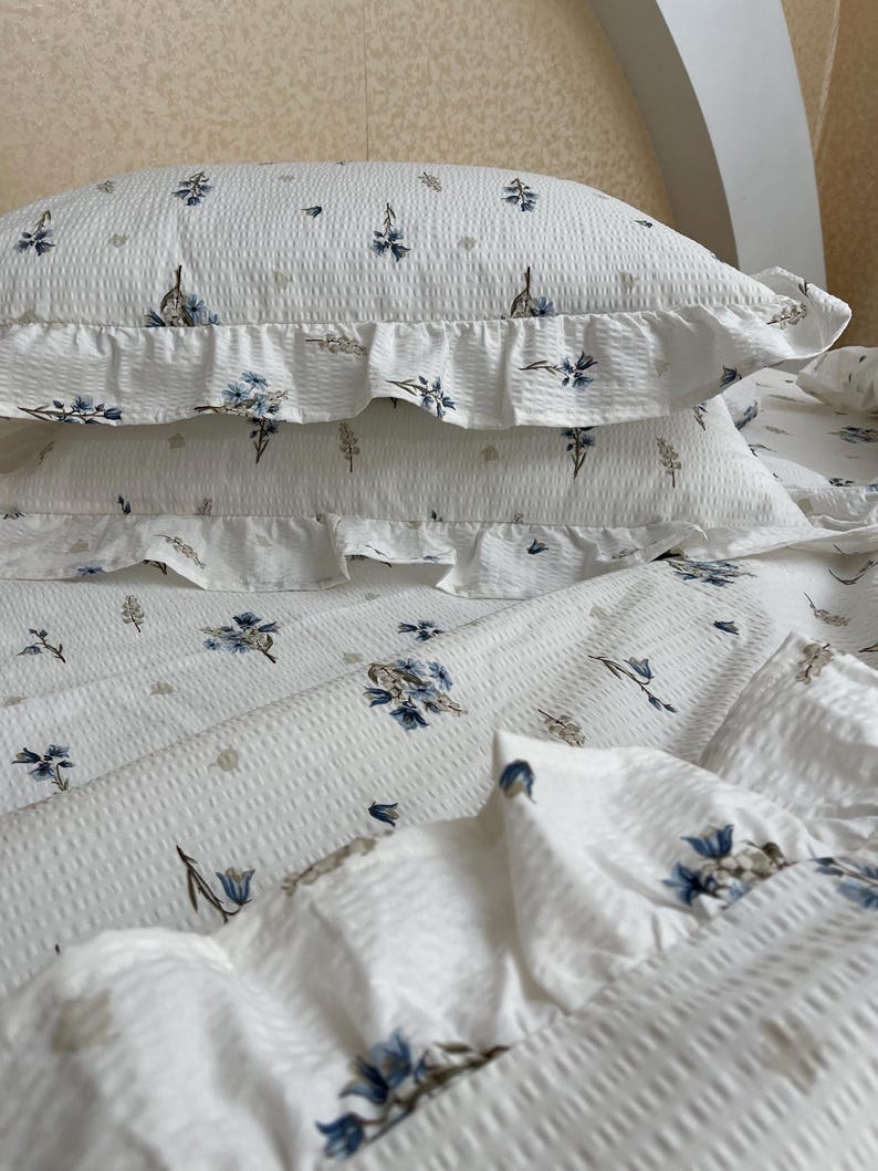 May include: White bedding set with a floral pattern in blue and beige. The set includes two ruffled pillowcases and a sheet, all with a textured, waffle-like weave. The floral design adds a touch of elegance to the bedroom decor.