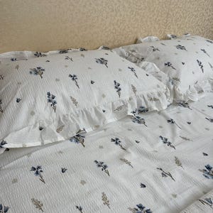 May include: White bedding set with a floral pattern in blue and gray. The set includes two pillowcases with ruffled edges and a matching sheet. The fabric has a textured, waffle-like appearance, adding visual interest to the design.