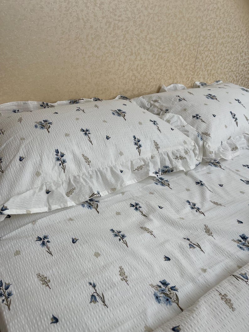 May include: White bedding set with a floral pattern in blue and gray. The set includes two pillowcases with ruffled edges and a matching sheet. The fabric has a textured, waffle-like appearance, adding visual interest to the design.