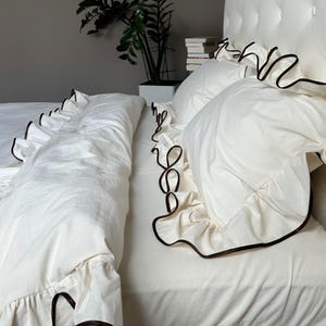 May include: A white bed set with ruffled edges and dark brown trim. The bedding includes a duvet cover and pillowcases. The headboard is white and tufted. A potted plant and books are in the background.