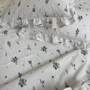 May include: White bedding set with a floral pattern in blue and gray. The set includes a pillowcase with a ruffled edge. The fabric has a textured, waffle-like appearance, and the floral design is scattered across the surface.