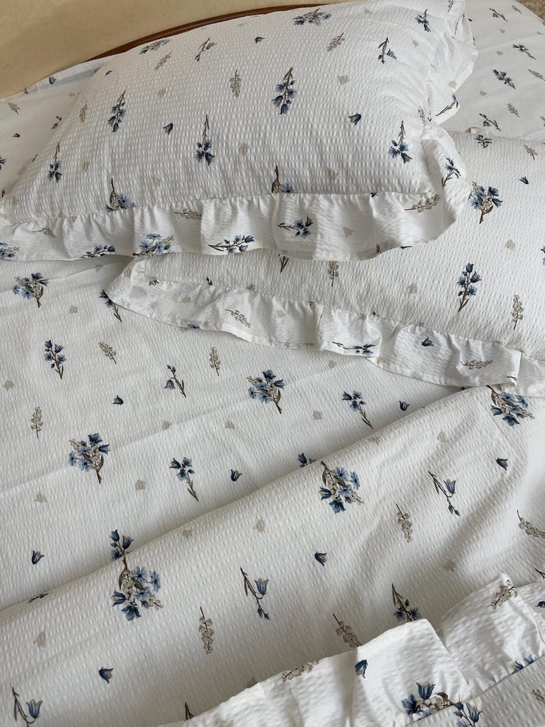May include: White bedding set with a floral pattern in blue and gray. The set includes a pillowcase with a ruffled edge. The fabric has a textured, waffle-like appearance, and the floral design is scattered across the surface.