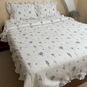 May include: White floral patterned bedding set featuring a quilt and two pillowcases. The quilt and pillowcases have a ruffled edge and are decorated with blue floral and leaf designs. The bedding is on a wooden bed frame.