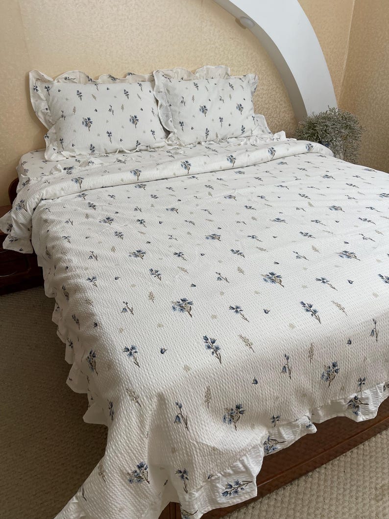 May include: White floral patterned bedding set featuring a quilt and two pillowcases. The quilt and pillowcases have a ruffled edge and are decorated with blue floral and leaf designs. The bedding is on a wooden bed frame.