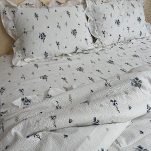 May include: White bedding set with a floral pattern. The set includes two ruffled pillowcases and a matching duvet cover. The floral design features blue and gray flowers and leaves, creating a soft, inviting aesthetic.