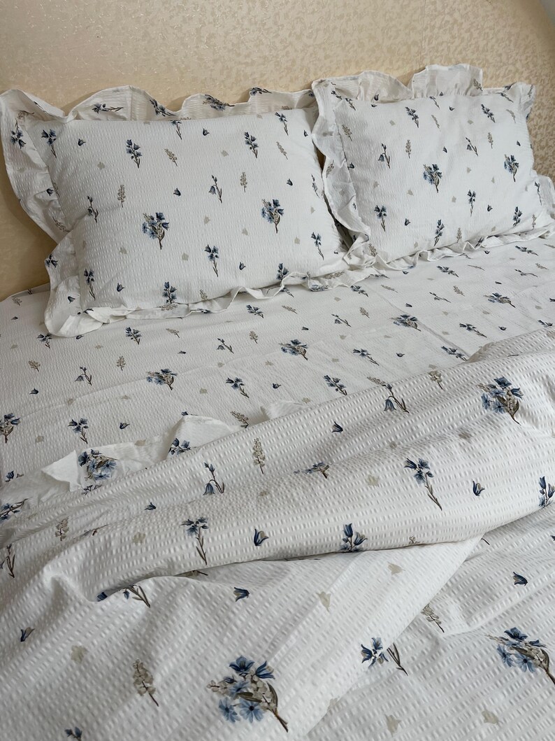 May include: White bedding set with a floral pattern. The set includes two ruffled pillowcases and a matching duvet cover. The floral design features blue and gray flowers and leaves, creating a soft, inviting aesthetic.