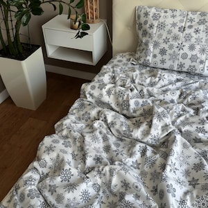 Könnte beinhalten: Weißes Bettwäsche-Set mit grauem Schneeflockenmuster. Das Set beinhaltet einen Kissenbezug und einen Bettbezug. Das Bett ist mit dem Bettwäsche-Set bezogen. Ein weißer Nachttisch und eine Topfpflanze befinden sich im Hintergrund.