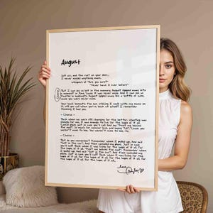 May include: A framed print featuring handwritten lyrics from the song "August." The text is in black ink on a white background, with the title "August" at the top. The frame is a light wood color, and the print is held by a person.