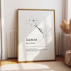 May include: A framed print with a minimalist design. The artwork features the word "Lumos" with a wand illustration and text describing the charm. The print is in a wooden frame and is displayed on a wall.