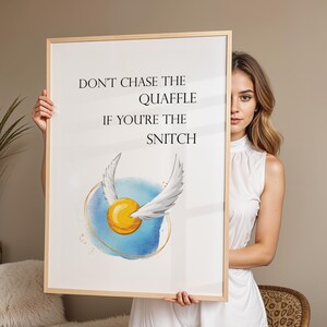 Puede incluir: Una impresión artística enmarcada con el texto "DON'T CHASE THE QUAFFLE IF YOU'RE THE SNITCH". La obra de arte incluye una snitch dorada con alas blancas sobre un fondo de acuarela azul y dorado. El marco es de color madera clara.