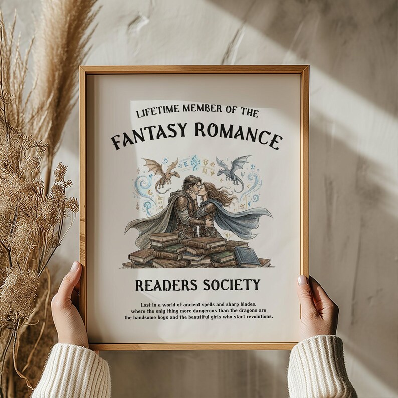 May include: A framed print with the text "LIFETIME MEMBER OF THE FANTASY ROMANCE READERS SOCIETY." The artwork depicts a couple kissing atop a stack of books, with dragons and decorative elements. The print has a gold frame.