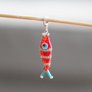 May include: A red and white beaded fish charm with a blue evil eye. The charm is attached to a silver lobster clasp.