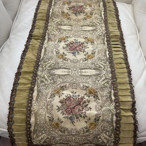 May include: A decorative table runner with a floral design. The runner has a cream-coloured background with floral medallions in pink, yellow, and blue. The edges are trimmed with green velvet and a decorative brown trim, adding a touch of elegance.