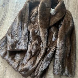 Könnte beinhalten: Ein brauner Pelzmantel mit breitem Kragen und langen Ärmeln. Der Mantel besteht aus einem weichen, strukturierten Material und scheint in gutem Zustand zu sein. Der Mantel liegt auf einer Holzoberfläche.