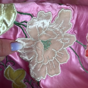 May include: Close-up of pink satin fabric with embroidered floral design. The embroidery features a large, light pink flower with white outlining and green accents. Additional yellow and green leaves are also visible.