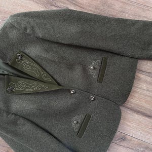 May include: A dark olive green wool-blend jacket featuring a high collar, long sleeves, and a button-front closure. The jacket includes dark green velvet lapel accents with white decorative embroidery and matching embroidered details above the pockets.