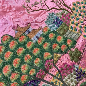 May include: Close-up of a pink embroidered textile featuring a detailed landscape scene. The design includes trees with green and orange foliage, a building with a brown roof, and various colorful plants. The embroidery is intricate and detailed.