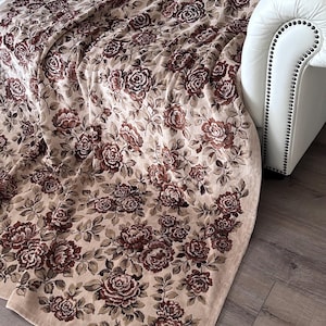 Könnte beinhalten: Eine beige Decke mit einem sich wiederholenden Muster aus burgunderroten Rosen und grünen Blättern. Die Decke ist über ein weißes Sofa drapiert, wobei ein Teil des Holzbodens sichtbar ist.