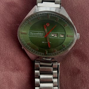 Puede incluir: Reloj de pulsera vintage plateado con esfera verde. El reloj tiene una pantalla de fecha y agujas rojas. La esfera del reloj tiene el texto "AbmonodZabod" y "CP". La correa del reloj es de metal plateado.