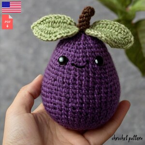 Puede incluir: Una higuera de ganchillo morada hecha a mano con una cara sonriente, ojos negros y hojas verdes. La higuera tiene un tallo marrón y se sostiene en una mano. El texto "chrochet pattern" es visible en la parte inferior.