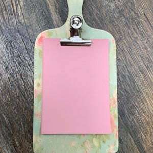 Può includere: Una lavagna portablocco decorativa con un motivo floreale verde chiaro e un inserto di carta rosa. Ha una clip in metallo argentato e un manico con un foro per appenderla. Il design è di ispirazione vintage.