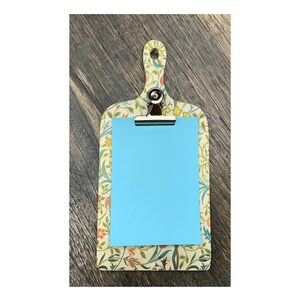 Può includere: Un portablocco decorativo con un motivo floreale e carta azzurra. Il portablocco ha una maniglia e una clip in metallo per tenere ferma la carta. Il motivo floreale comprende fiori e foglie verdi, arancioni e gialle.