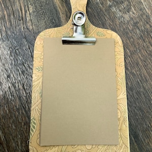 Può includere: Una lavagna portablocco decorativa con manico in legno e clip in metallo. La lavagna presenta un motivo floreale nei toni del beige, verde e pesca. Un foglio di carta bianco, color marrone chiaro, è tenuto in posizione dalla clip.