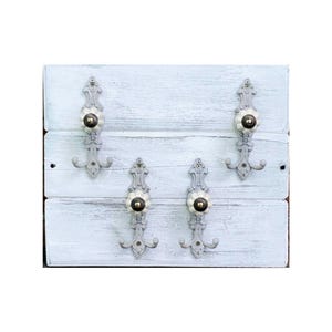 Pode incluir: Um cabide de parede de madeira branca desgastada com quatro ganchos de metal cinza ornamentados. Cada gancho apresenta um botão floral decorativo com um centro creme. O cabide é feito de tábuas de madeira horizontais.
