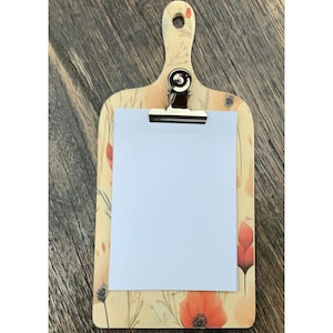 Può includere: Un portablocco decorativo con un design floreale con papaveri rossi e fogliame verde. Il portablocco ha un manico in legno e una clip in metallo che tiene un foglio di carta bianco vuoto. Lo sfondo è una superficie in legno.