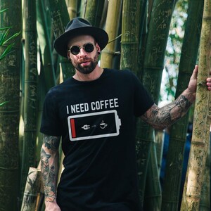Puede incluir: Un hombre con un sombrero negro y gafas de sol se encuentra frente a un bosque de bambú. Lleva una camiseta negra con el texto "I NEED COFFEE" y un gráfico de una batería con un icono de taza de café.
