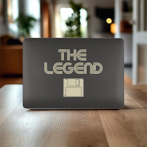 Op de afbeelding: Een donkergrijze laptop met de tekst "THE LEGEND" in een retro lettertype en een floppy disk-pictogram. De laptop staat op een houten tafel, met een vage achtergrond.