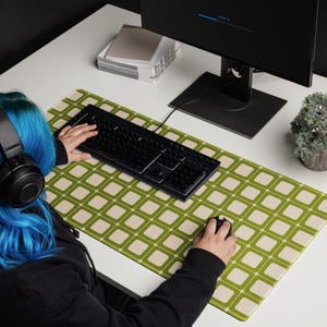 Op de afbeelding: Een rechthoekige computermuispad met een retro geometrisch patroon van olijfgroene vierkanten op een crèmekleurige achtergrond. Een zwart toetsenbord en muis liggen op de pad. De pad ligt op een witte tafel met een computermonitor en een kleine potplant.