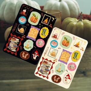 May include: Two sticker sheets with autumn illustrations. One is black, the other cream. Each features a pumpkin, sweater, books, and other fall images. The background includes pumpkins and a candle.