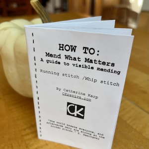 May include: A white booklet titled "HOW TO: Mend What Matters" with text about visible mending, next to a small white pumpkin. The booklet includes instructions for running and whip stitches, and the author's name.