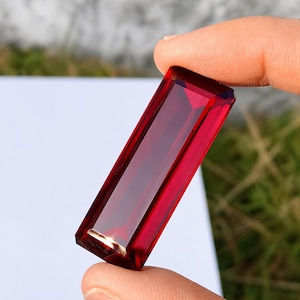 May include: A rectangular, faceted, deep red gemstone is held between fingers. The gem has a polished surface and a rich, transparent color. The cut of the stone allows light to pass through, creating a brilliant display. The gemstone is a striking example of fine jewelry.