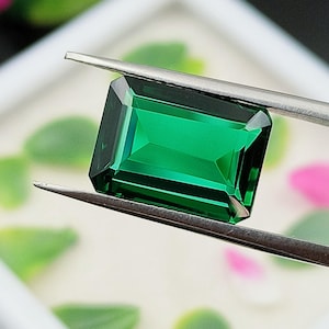 May include: A vibrant emerald green gemstone with a rectangular emerald cut, held by a pair of silver-toned metal tweezers. The gemstone features sharp, clean facets that reflect light, showcasing a deep, saturated green colour throughout the stone.
