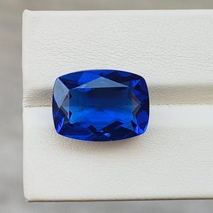 Könnte beinhalten: Ein lebendiger, kissenförmig geschliffener Saphir-Edelstein. Der tiefblaue Stein ist vor einem weißen Hintergrund platziert, der seine Facetten und seinen Glanz hervorhebt. Die Form des Edelsteins ist rechteckig mit abgerundeten Ecken.