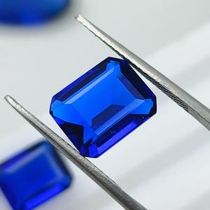Pode incluir: Uma gema retangular de um azul profundo e vibrante com facetas de corte esmeralda, segurada por uma pinça de metal prateado. Duas gemas azuis adicionais, uma retangular e uma oval, são visíveis no fundo desfocado, exibindo cor saturada.