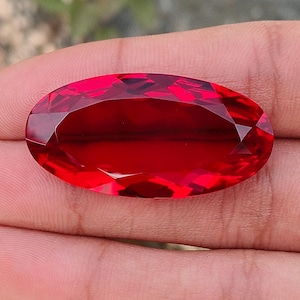 Pode incluir: Uma gema vermelha escura facetada, em forma oval. A gema é segurada em uma mão, com as facetas captando a luz. A cor da pedra é um vermelho rico e vibrante, e o corte cria um efeito brilhante. O fundo está desfocado, com folhagem verde.