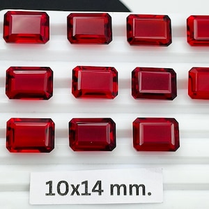 May include: Eleven rectangular red gemstones with faceted surfaces arranged in three rows on a white grooved display stand. A white rectangular label at the bottom center displays the text 10x14 mm. indicating the size of the individual stones.