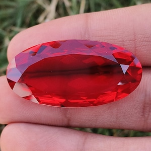 Könnte beinhalten: Ein facettierter, oval geformter, tiefroter Edelstein. Der Edelstein wird in einer Hand gehalten, wodurch sein brillanter Schliff und seine satte Farbe zur Geltung kommen. Die Facetten des Steins fangen das Licht ein und erzeugen einen funkelnden Effekt.