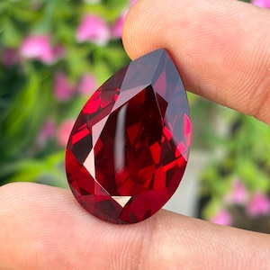 May include: A pear-shaped, faceted gemstone with a deep, vibrant red color. The stone features a polished surface with multiple geometric facets that reflect light, creating a brilliant appearance with sharp edges and a pointed top.