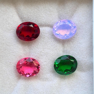 Puede incluir: Cuatro piedras preciosas talladas en forma ovalada dispuestas en una caja expositora blanca. La colección incluye una piedra roja oscura, una lavanda pálida, una rosa vibrante y una verde esmeralda, sobre un forro de terciopelo claro.