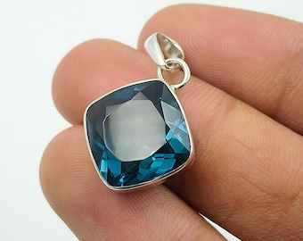 May include: A square-cut, blue gemstone pendant with a silver-colored setting. The gemstone has a faceted surface, reflecting light. The pendant is suspended from a small loop, ready for a chain. The background is plain white.