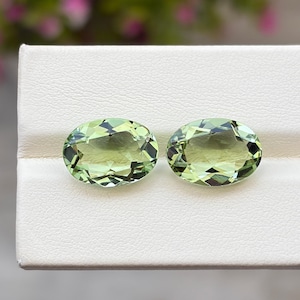 May include: Two oval-shaped, faceted light green gemstones displayed side by side. The stones feature a transparent, pale lime-green hue with intricate geometric facets that reflect light across their polished, symmetrical surfaces.