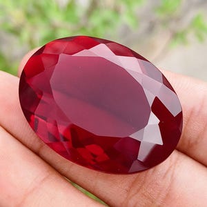 Puede incluir: Una gema grande, facetada, de forma ovalada y color rojo intenso. La piedra preciosa se sostiene en una mano, con un fondo verde borroso. Las facetas de la piedra capturan la luz, creando reflejos y sombras. La gema parece ser un rubí.