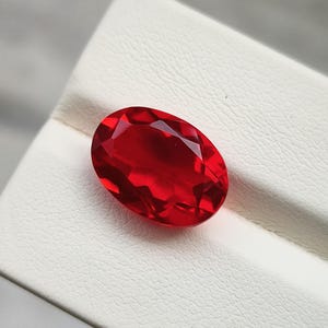 Peut inclure: Une pierre précieuse rouge vif, de forme ovale et facettée. La gemme est posée sur une surface blanche texturée, la lumière se reflétant sur ses nombreuses facettes. Un bijou captivant.
