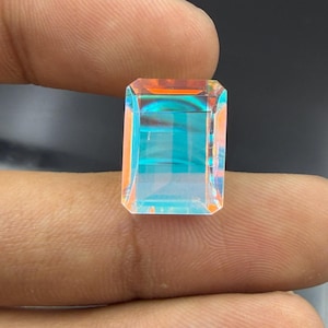 Peut inclure: Une pierre précieuse rectangulaire à facettes avec une finition irisée, affichant un jeu de lumière vibrant avec des teintes changeantes de bleu turquoise vif et de tons orange-rose sur toute la surface transparente de la pierre.