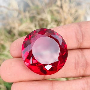 Pode incluir: Uma grande gema redonda, facetada, vermelho escuro. A gema é segurada em uma mão, mostrando seu corte e cor brilhantes. As facetas da gema refletem a luz, criando um efeito brilhante. O fundo está desfocado.